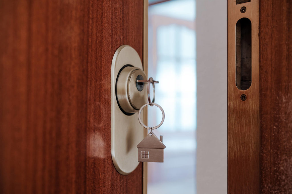 Foto da porta entreaberta, com uma chave e um chaveiro no formato de casa. Imagem para ilustrar o texto sobre padronização na vistoria de imóveis
